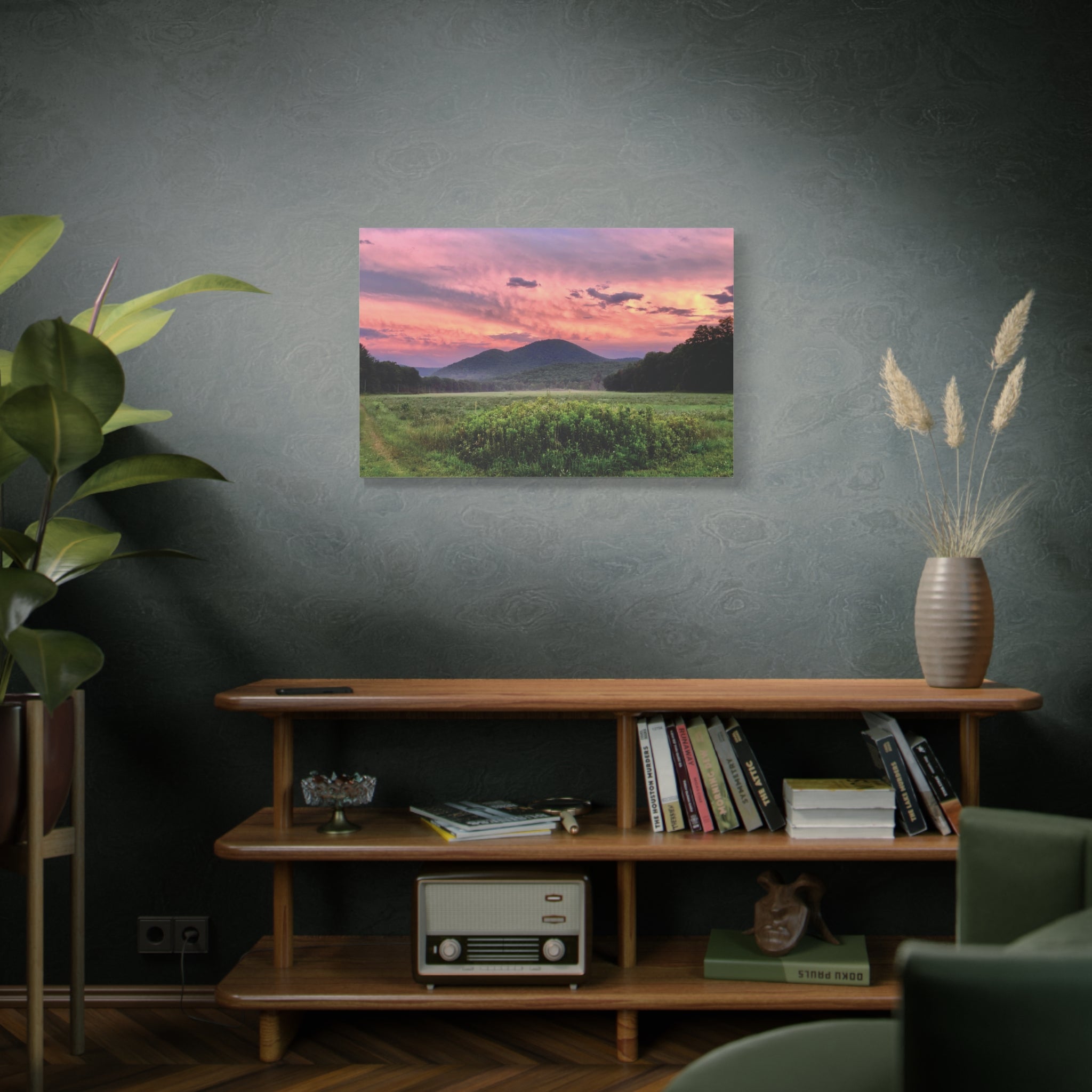 Lavender sunset over a field with mountains, displayed on a wall above a wooden shelf.